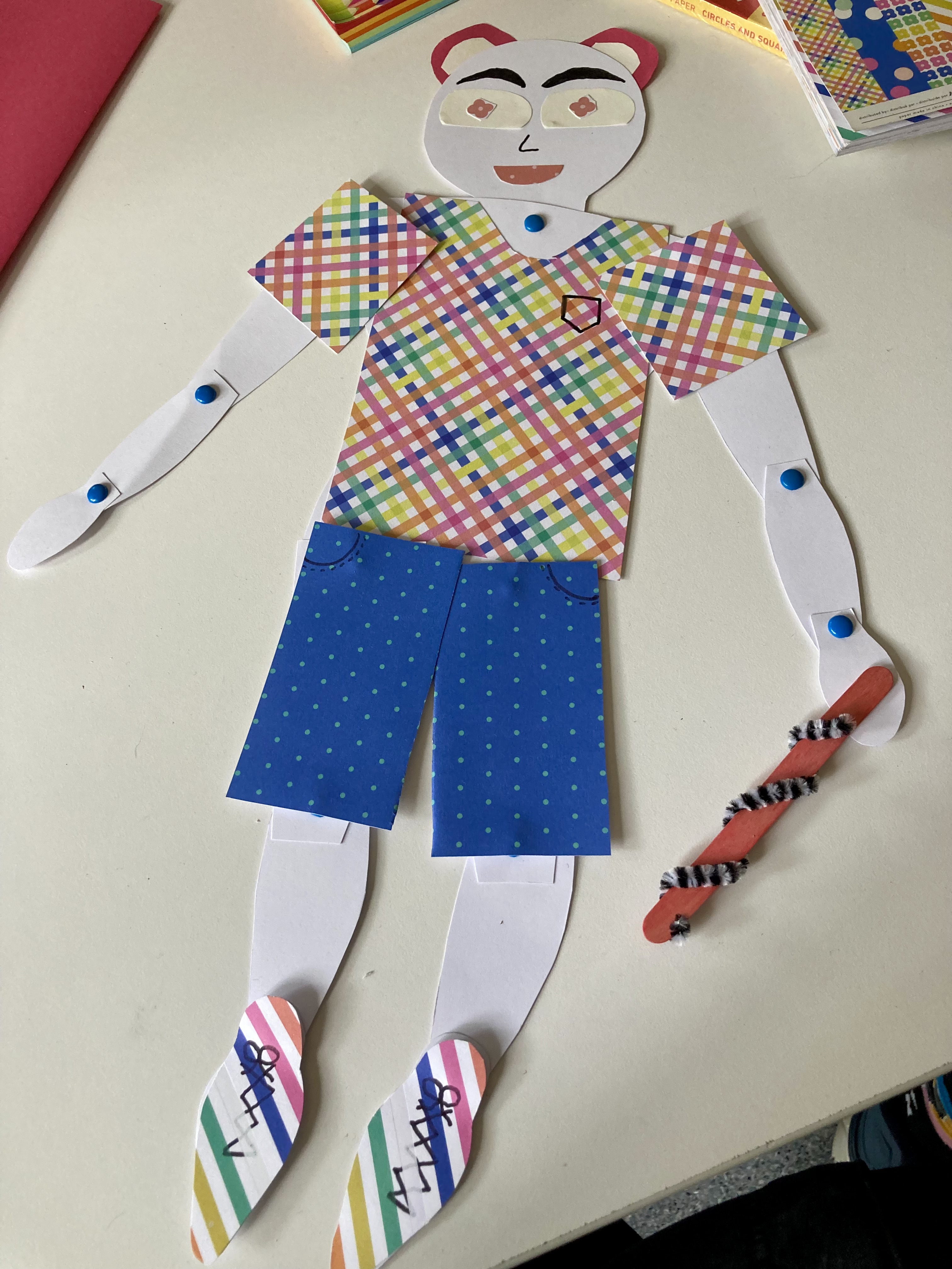 A jointed puppet wearing a rainbow plaid shirt, blue spotty shorts and a pair of pink bear ears. They are carrying a walking stick make out of a popsicle stick with a pipecleaner wrapped around it.