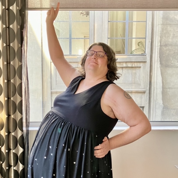 A woman wearing a fancy black dress standing in front of a window, one hand on her hip and the other pointing upwards